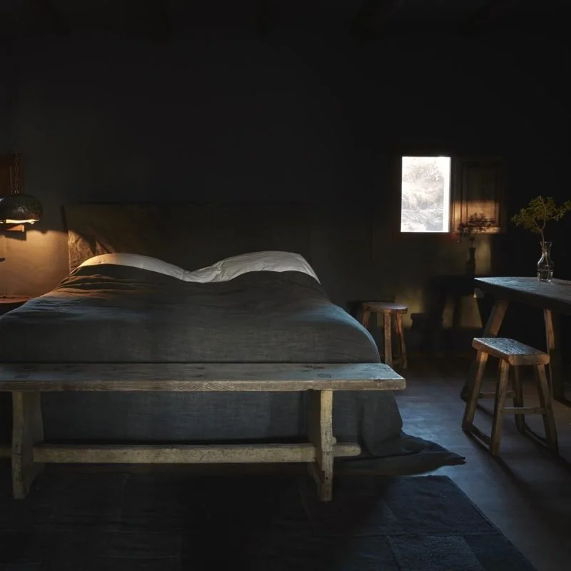 A dark bedroom with a bed, a window, and some furniture, illuminated by natural light coming through the window.