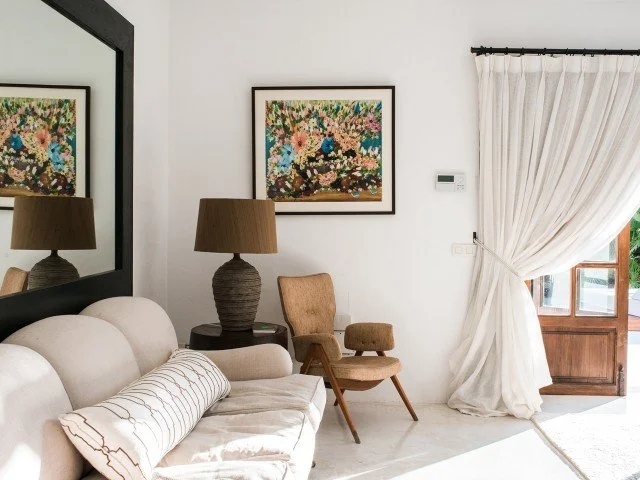 Living room with a beige sofa, brown table lamps, a wooden chair, framed floral artwork, and white curtains over a window