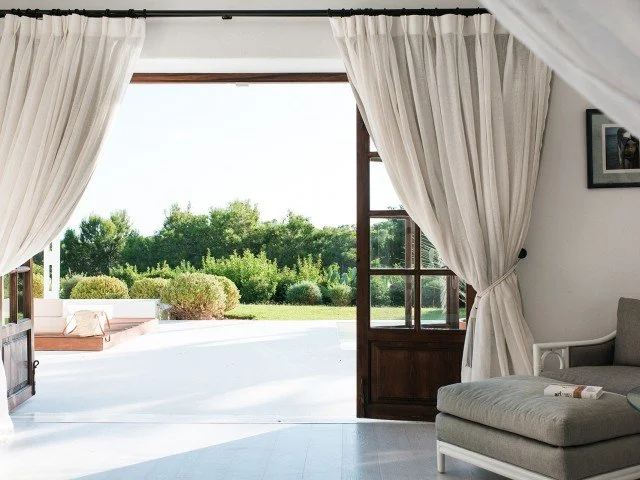 Open door leading to a bright outdoor patio and garden with green bushes and trees, white curtains, and a gray sofa inside a room.