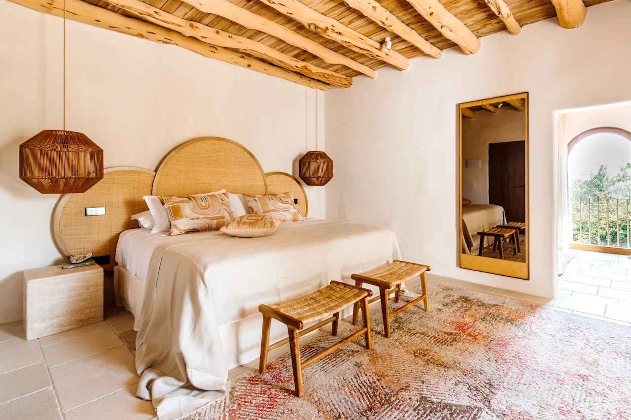 A bedroom with a large bed, wooden accents, and natural light, featuring rattan pendant lights, a large headboard, and a mirror reflecting part of the room.