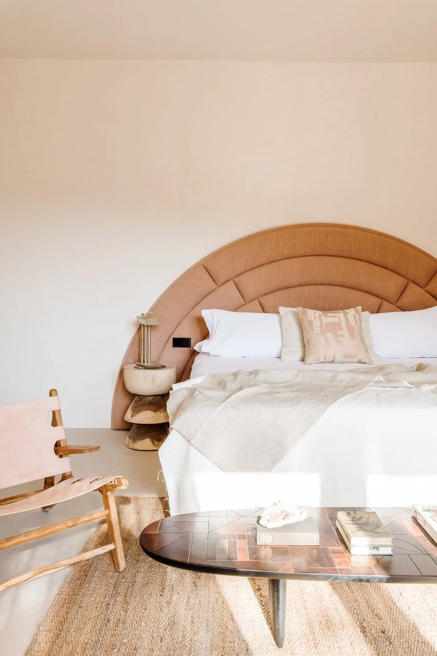 A bedroom with a beige and pink color scheme, featuring a large bed with white linens, beige throw blanket, and decorative pillows, a unique half-circle upholstered headboard, a wooden chair with a leather seat, a dark wood coffee table with books an