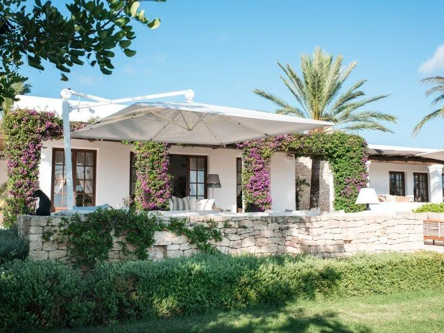 A white villa with purple flowering vines, outdoor sofa, and umbrellas on a sunny day.