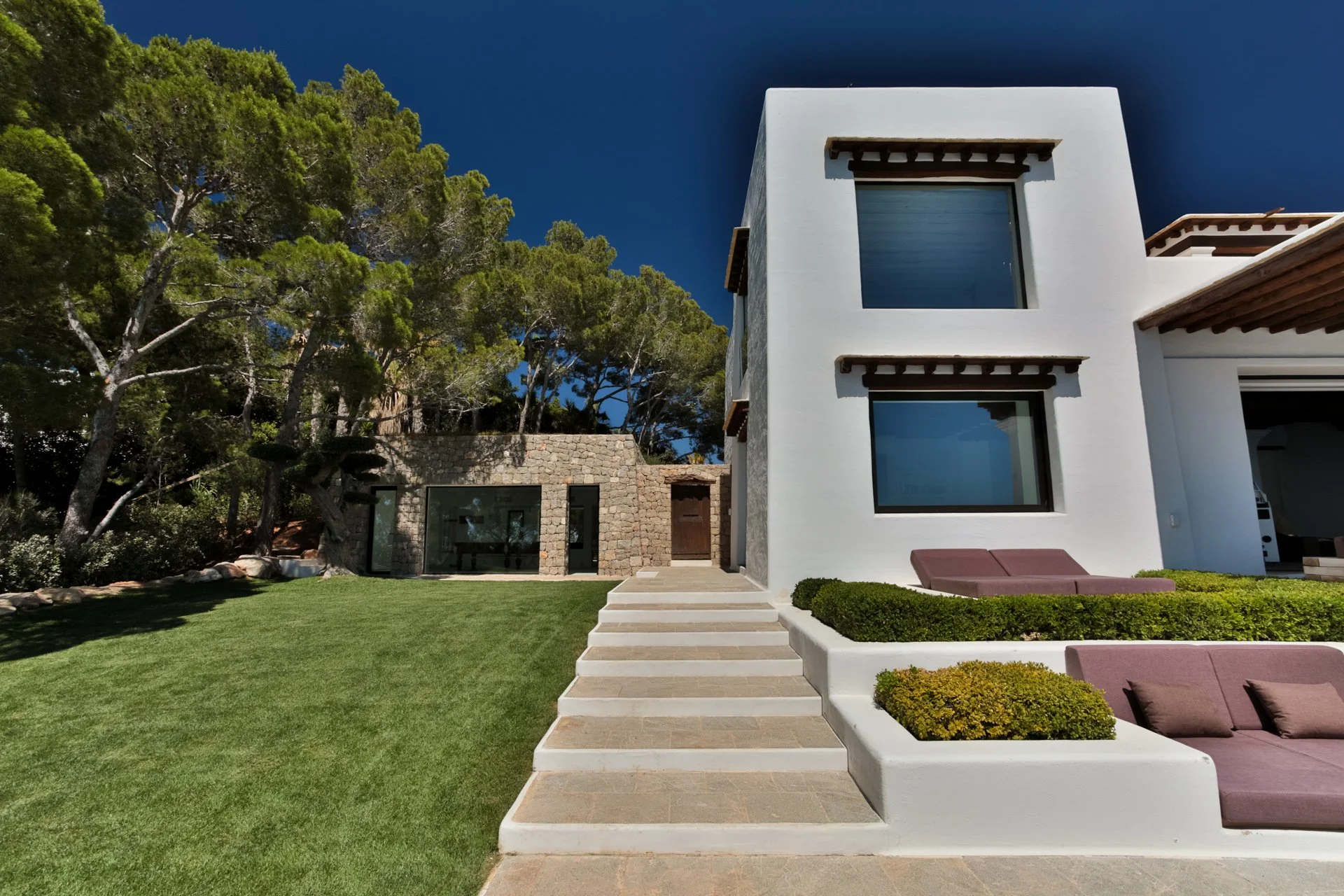 Modern two-story house with white exterior, large windows, and wooden accents, surrounded by a green lawn and trees, under a clear blue sky.