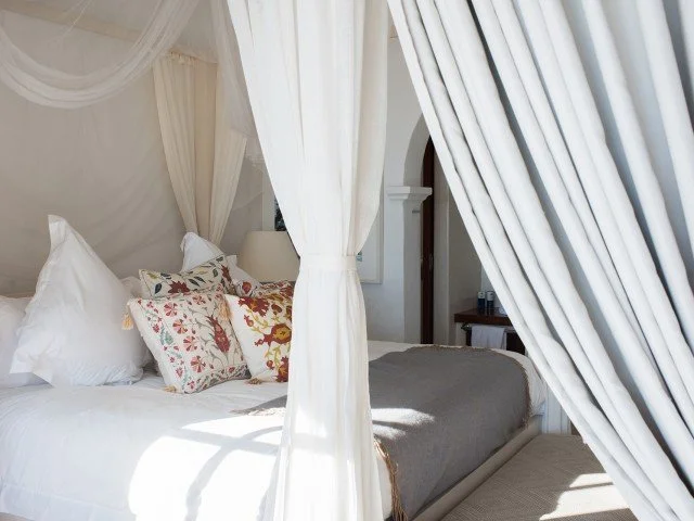 A cozy bedroom with a four-poster bed, white bedding and pillows, some with floral and animal designs, and sheer white curtains.