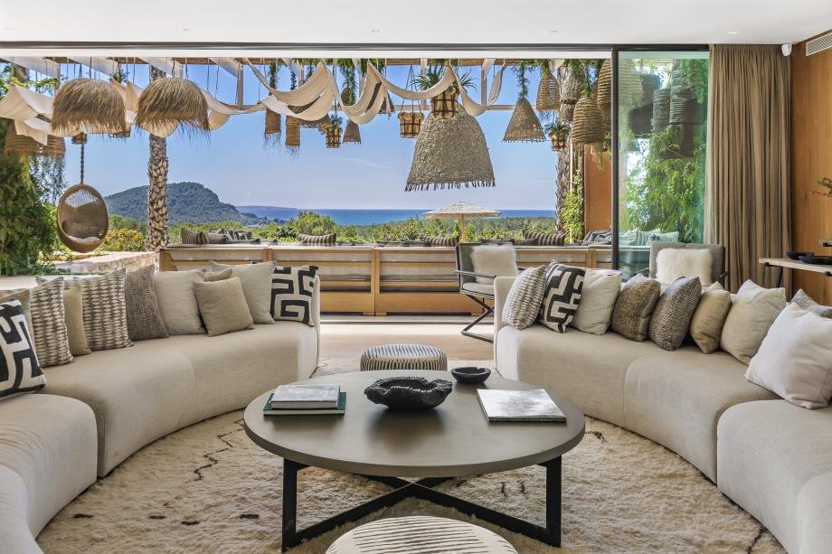 Living room with large open window showing a tropical landscape and ocean view, decorated with hanging wicker and fabric art pieces, beige sofas with patterned cushions, round coffee table with books and decorative objects, and a cozy, modern aesthet