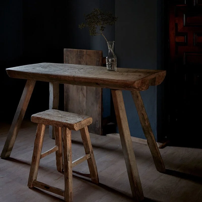 A rustic wooden table and stool with a glass vase containing a single branch against a dark wall.