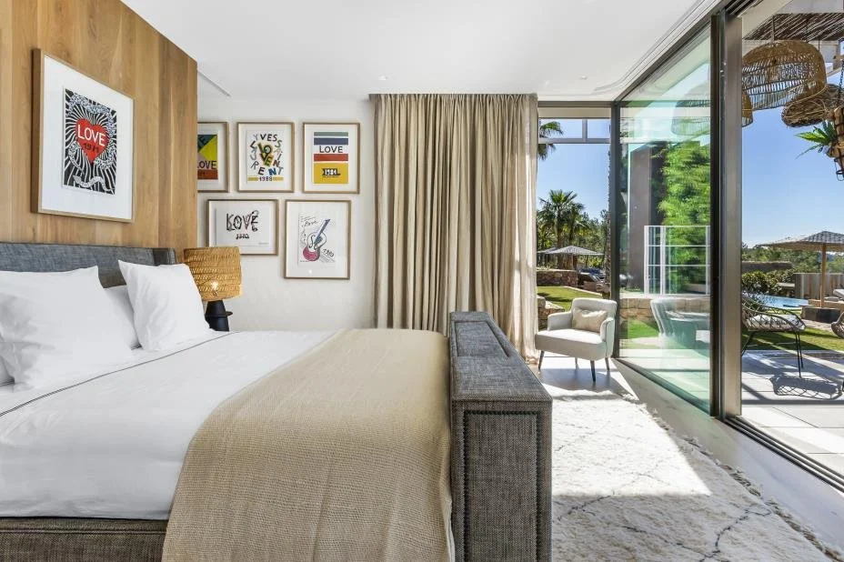 A modern bedroom with a bed, framed artwork on the wall, a bedside lamp, and large glass sliding doors leading to an outdoor patio with lounge chairs, umbrellas, and palm trees.