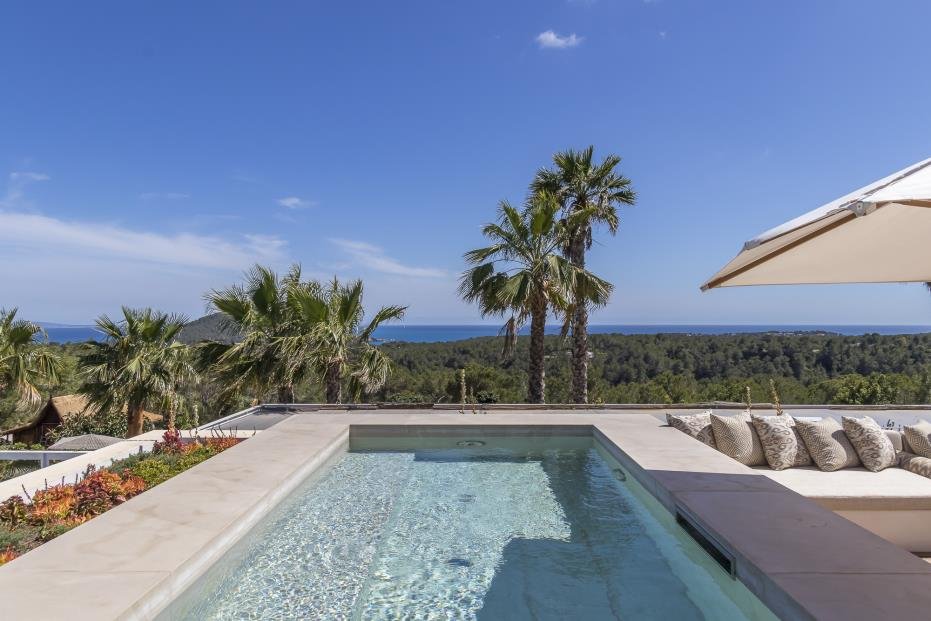 Poolside view with lounge chairs, palm trees, and ocean in the background under blue sky.