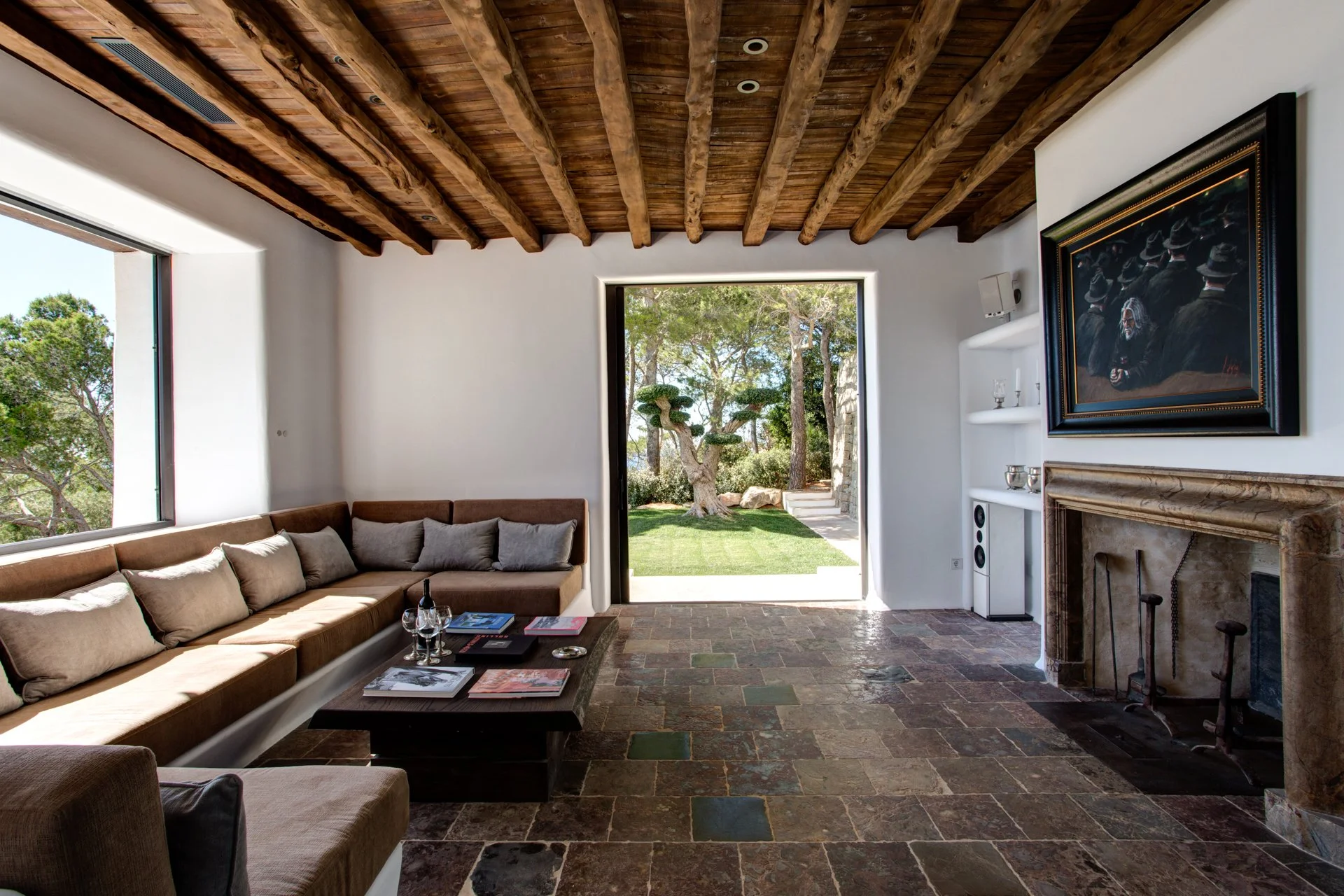 Living room with a curved sofa, stone floor, wooden ceiling beams, fireplace, large open doorway showing a garden with trees