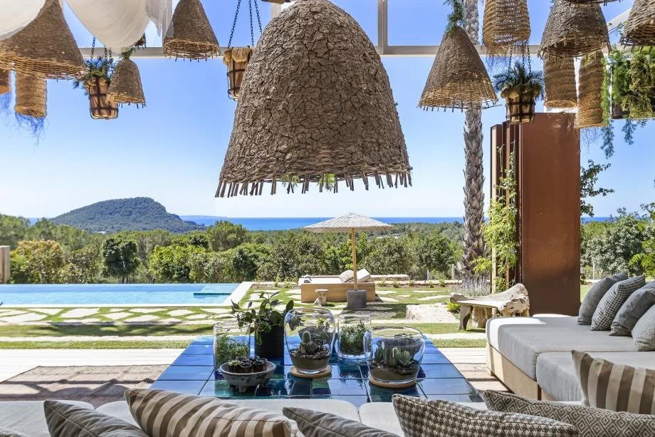 Living room with a view of the outdoors, including a swimming pool, trees, and ocean in the distance. The room features hanging woven light fixtures, a gray sofa with cushions, a coffee table with plants and decor, and large windows.