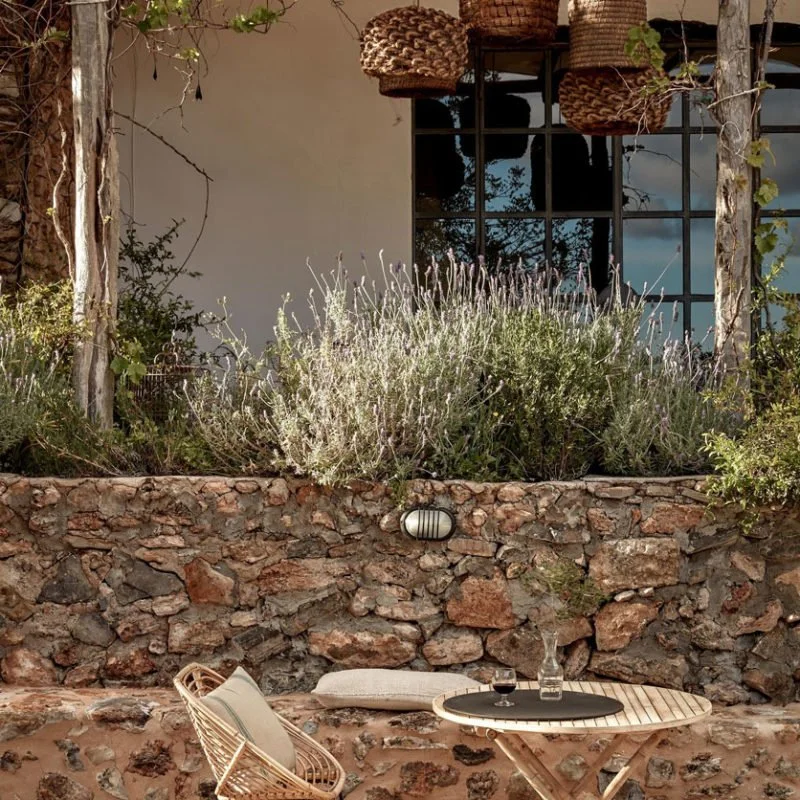 Outdoor patio with a round wooden table set with a wine glass and a water bottle, an outdoor chair with cushions, a stone retaining wall, lush greenery, and a window with hanging wicker baskets above.