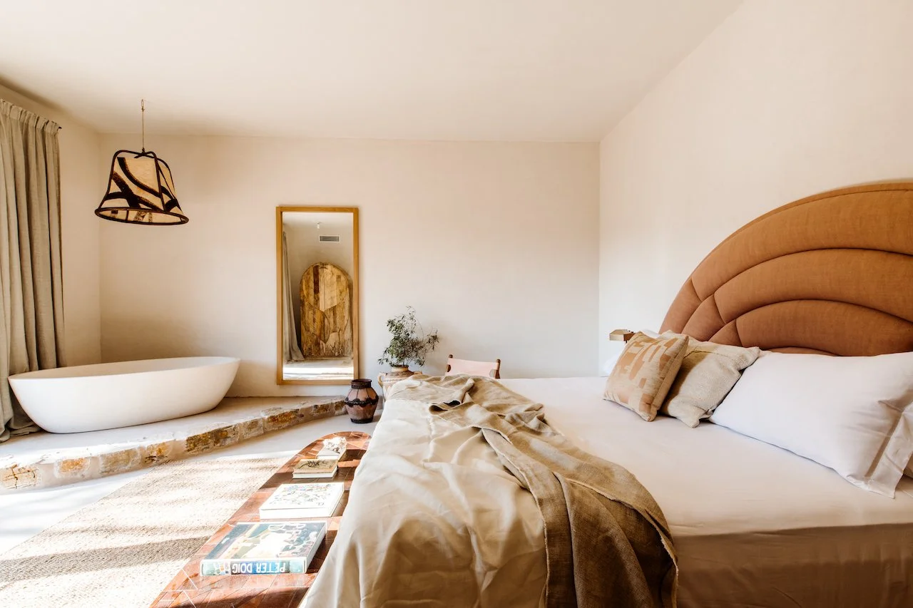 A cozy bedroom with a large bed, beige headboard, and pillows; a side table with plants and decor; a large mirror on the wall; a free-standing bathtub near a large window with curtains; a hanging lamp; books on a wooden coffee table; and a rustic bri