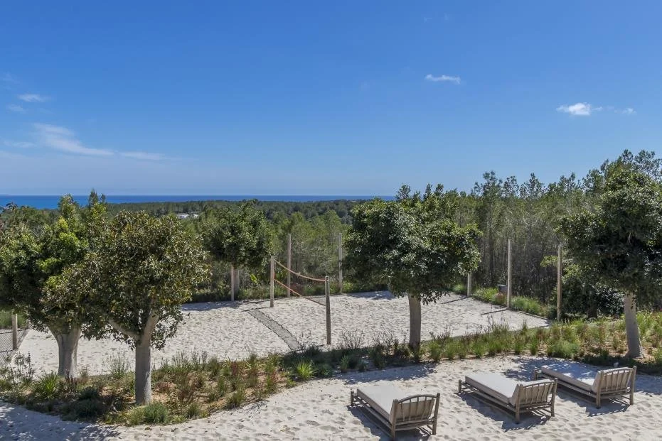 Beach area with sand, outdoor benches, and trees, overlooking the ocean under a blue sky.