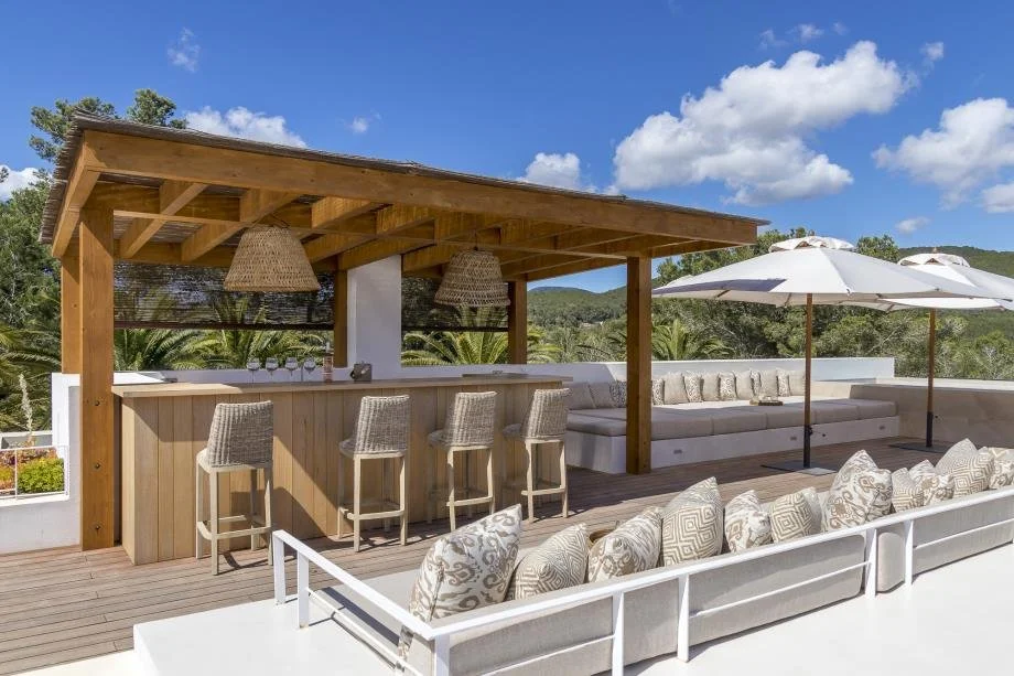 An outdoor rooftop bar area with a wooden pergola, woven pendant lights, a long wooden bar counter with four bar stools, and a seating area with plush lounge chairs, pillows, and large white umbrellas, overlooking a green landscape and blue sky.