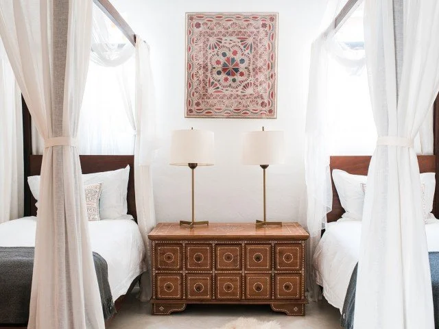 A bedroom with two twin beds separated by a wooden nightstand with two lamps, white curtains, and a colorful framed artwork hanging on the wall.