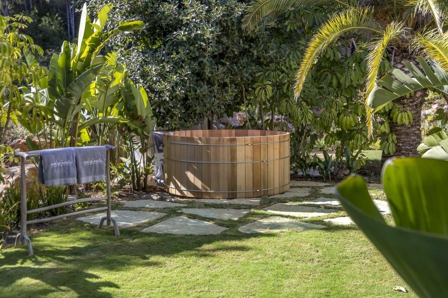 Wooden hot tub in a lush backyard garden surrounded by tropical plants and trees.