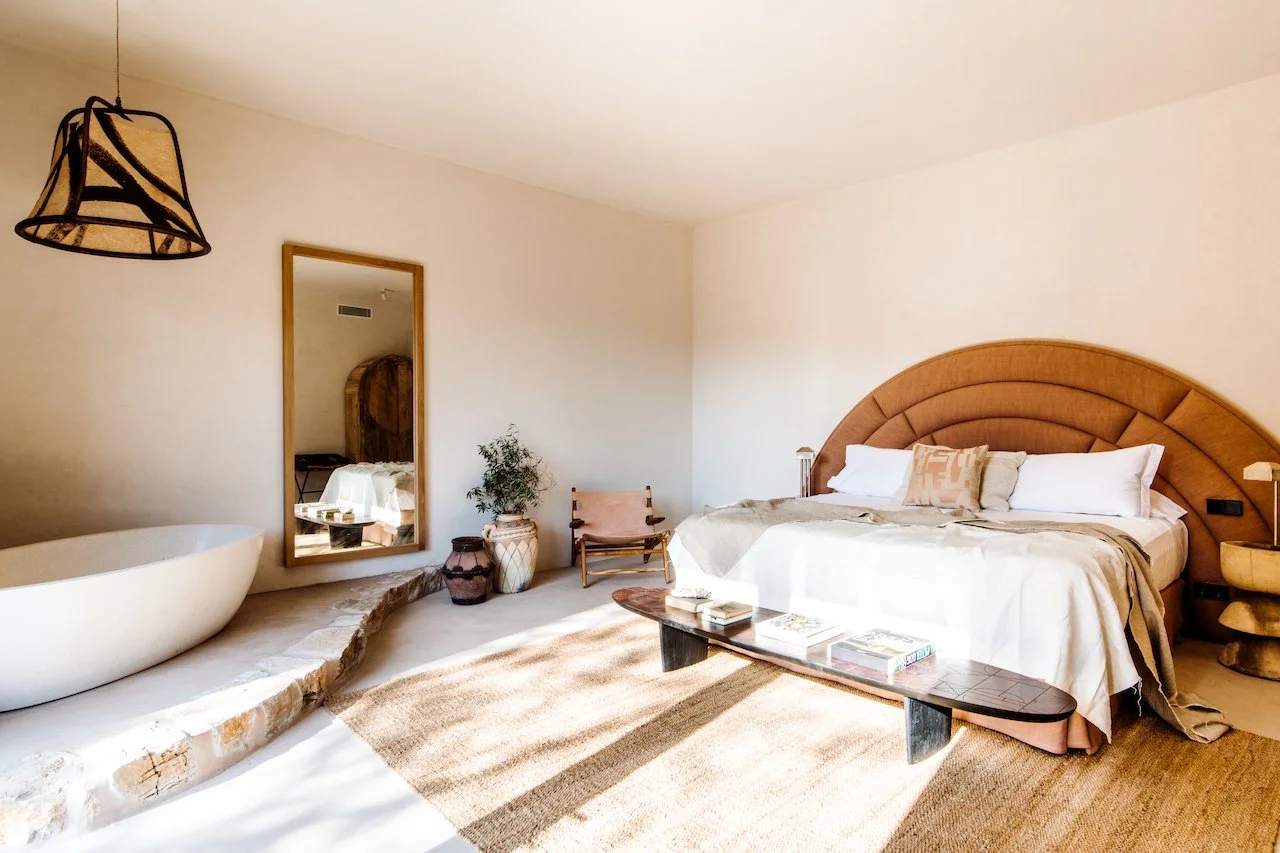 Minimalist bedroom with a large bed featuring an arched headboard, a wooden bench at the foot of the bed, a mirror on the wall, a small table, and decorative potted plants.