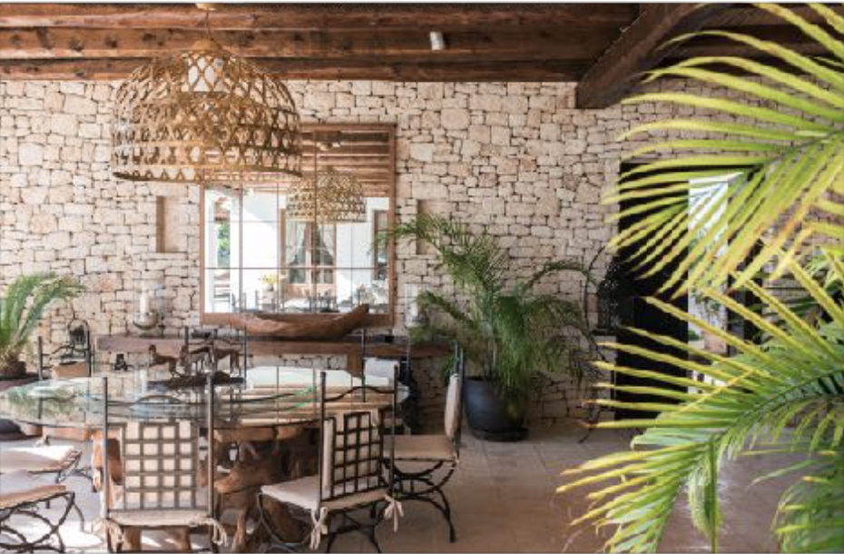 Rustic dining room with exposed brick walls, wooden ceiling beams, wicker pendant lights, a glass-top table surrounded by mixed chairs, a large mirror on the wall, and potted tropical plants.