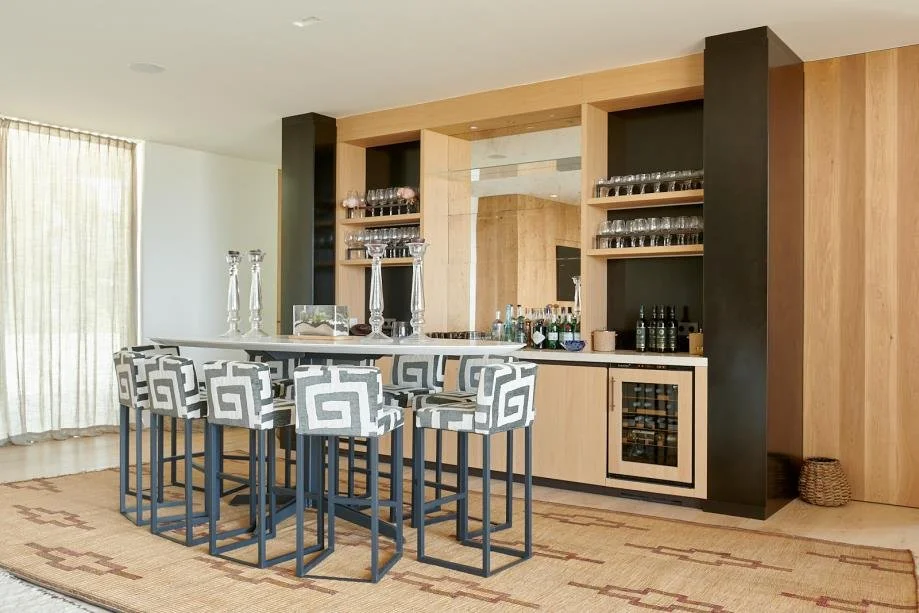Modern home bar with patterned bar stools, a white countertop, glassware on shelves, and a built-in wine fridge, set against wooden and black accents.