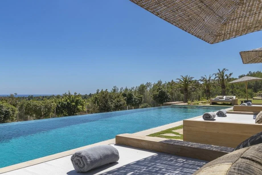 View of a luxurious outdoor infinity pool with rolled towels on the edge, surrounded by lounge chairs with umbrellas, overlooking lush greenery and a distant ocean under a clear blue sky.