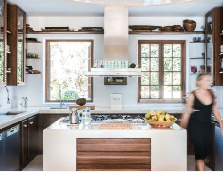 Modern kitchen with wooden shelves, white walls, and large windows, featuring a white island with a bowl of apples, and a woman in a black dress walking past.