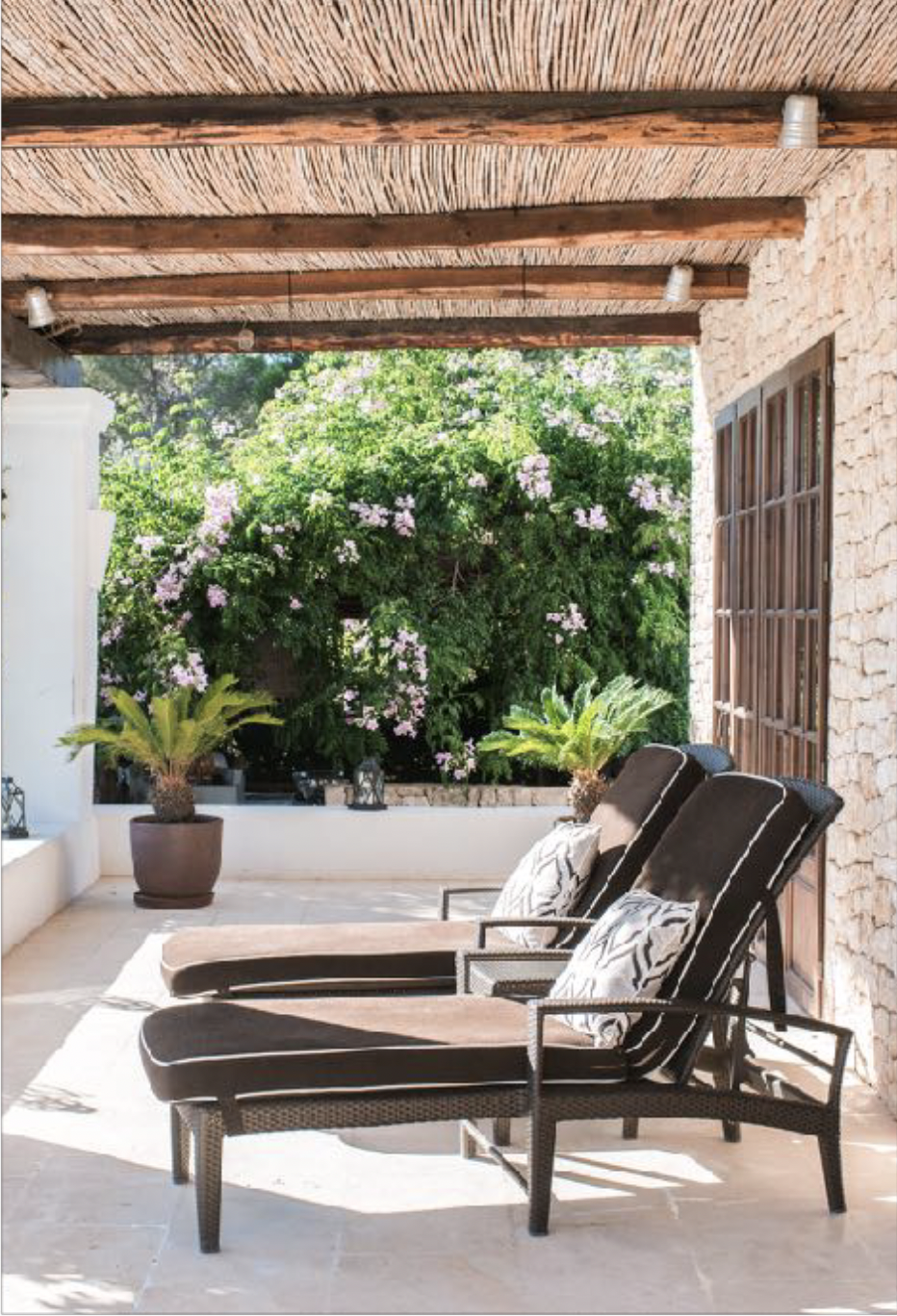 Outdoor patio with two lounge chairs and pillows, potted plants, a flowering bush in the background, and a wooden ceiling.