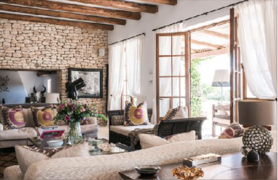 Living room with beige sofas, colorful cushions, a brick fireplace, wooden ceiling beams, and large windows with white curtains, opening to a balcony.