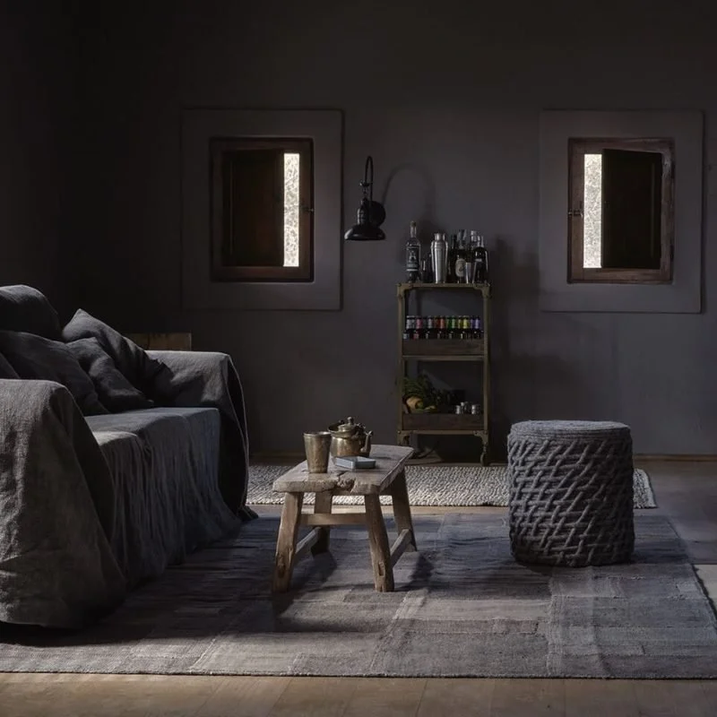 A cozy living room with dark walls, two small windows, a dark sofa with cushions, a wooden coffee table with metal cups and a teapot, a woven pouf, and a small shelf with bottles and spices, lit by natural light from the windows.