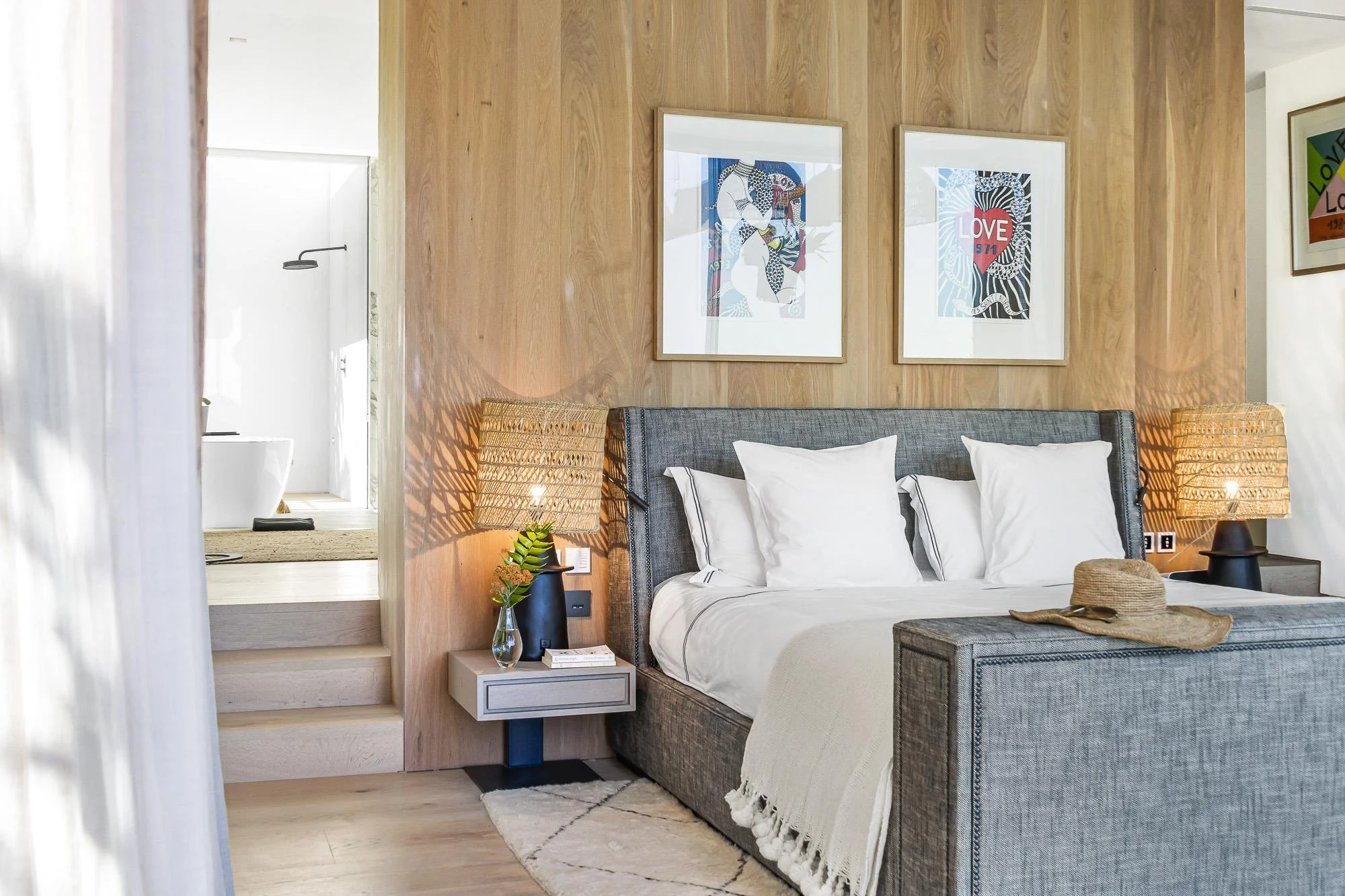 Modern bedroom with wooden accent wall, framed artwork, gray upholstered bed with white bedding, wicker lamps on nightstands, and a straw hat on a bench.