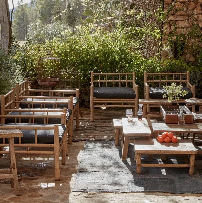Outdoor patio with wooden furniture, black cushioned chairs, small tables, plants, and a stone wall.