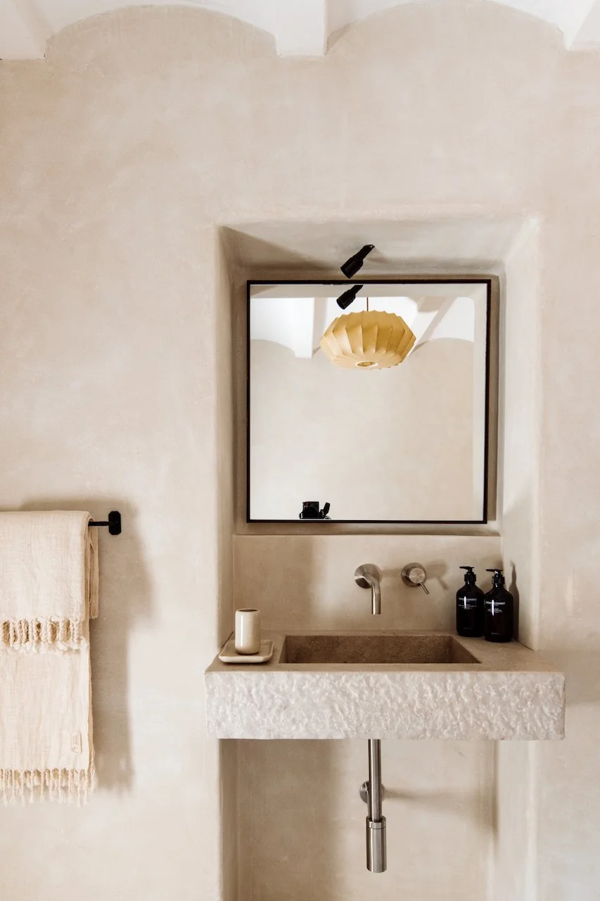 A minimalist bathroom with a textured stone sink, a wall-mounted faucet, a mirror reflecting a hanging beige pendant light, and black pump bottles on the side of the sink.