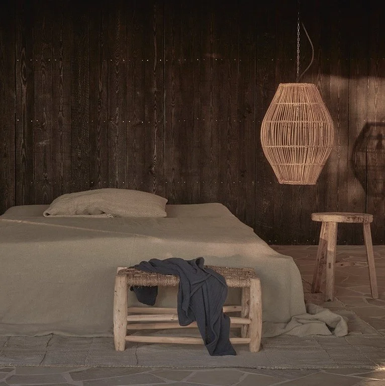 A rustic bedroom with a bed covered in a neutral-colored blanket and pillow, a small wooden bench at the foot of the bed with a dark clothing item draped over it, a wooden wall in the background, a hanging wicker light fixture, and a small wooden sto