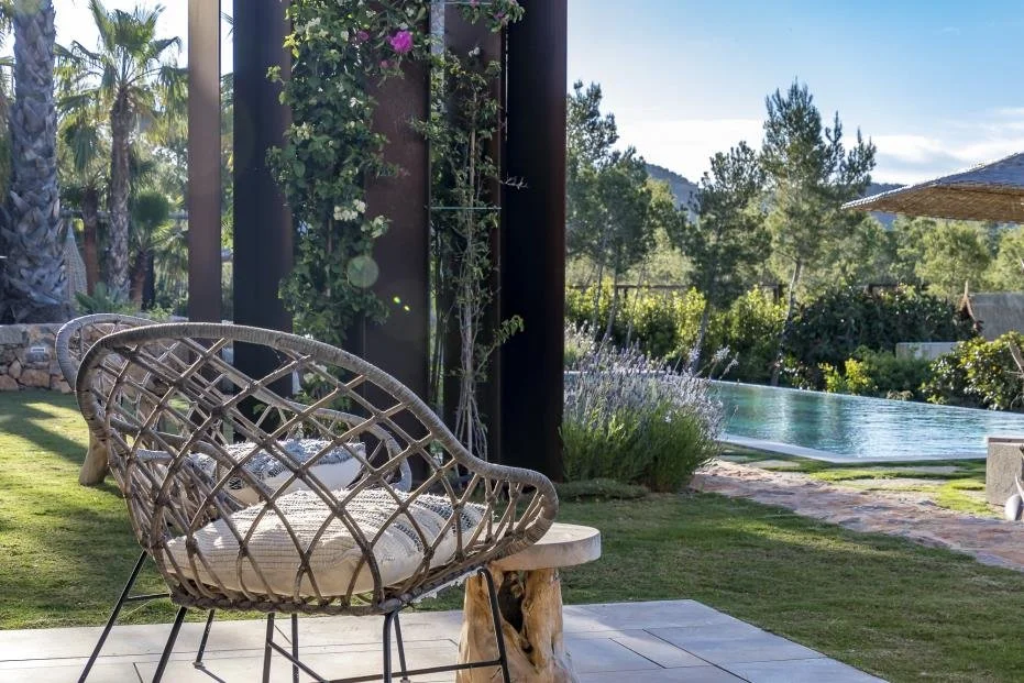 Outdoor patio with two wicker chairs and a small wooden table beside a pool, surrounded by lush greenery and trees under a clear blue sky.