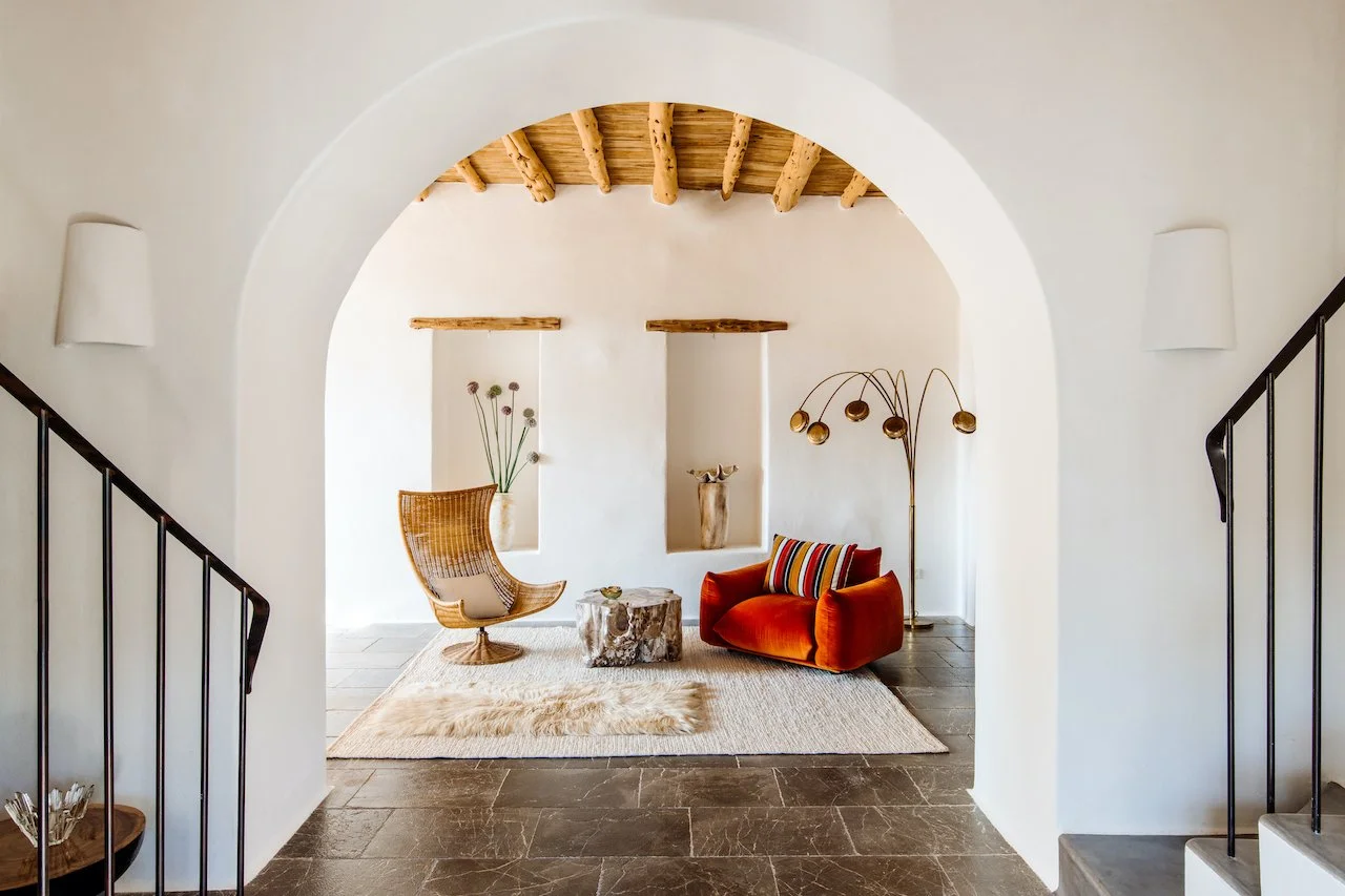 Living room with white walls, arched entrance, wooden ceiling beams, orange sofa with striped pillow, rattan armchair, modern floor lamp, decorative vases, and grassy wall art, with tiled floor and minimalist decor.