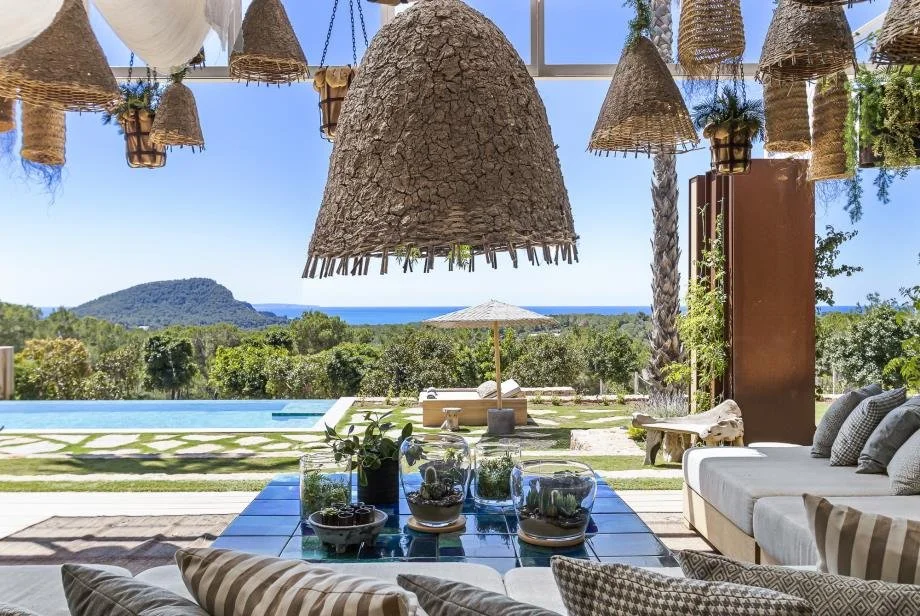 A bright indoor living area with large woven pendant lights hanging from the ceiling, overlooking a backyard with a pool, lounge chairs, and umbrellas, with a view of a distant hill and ocean.