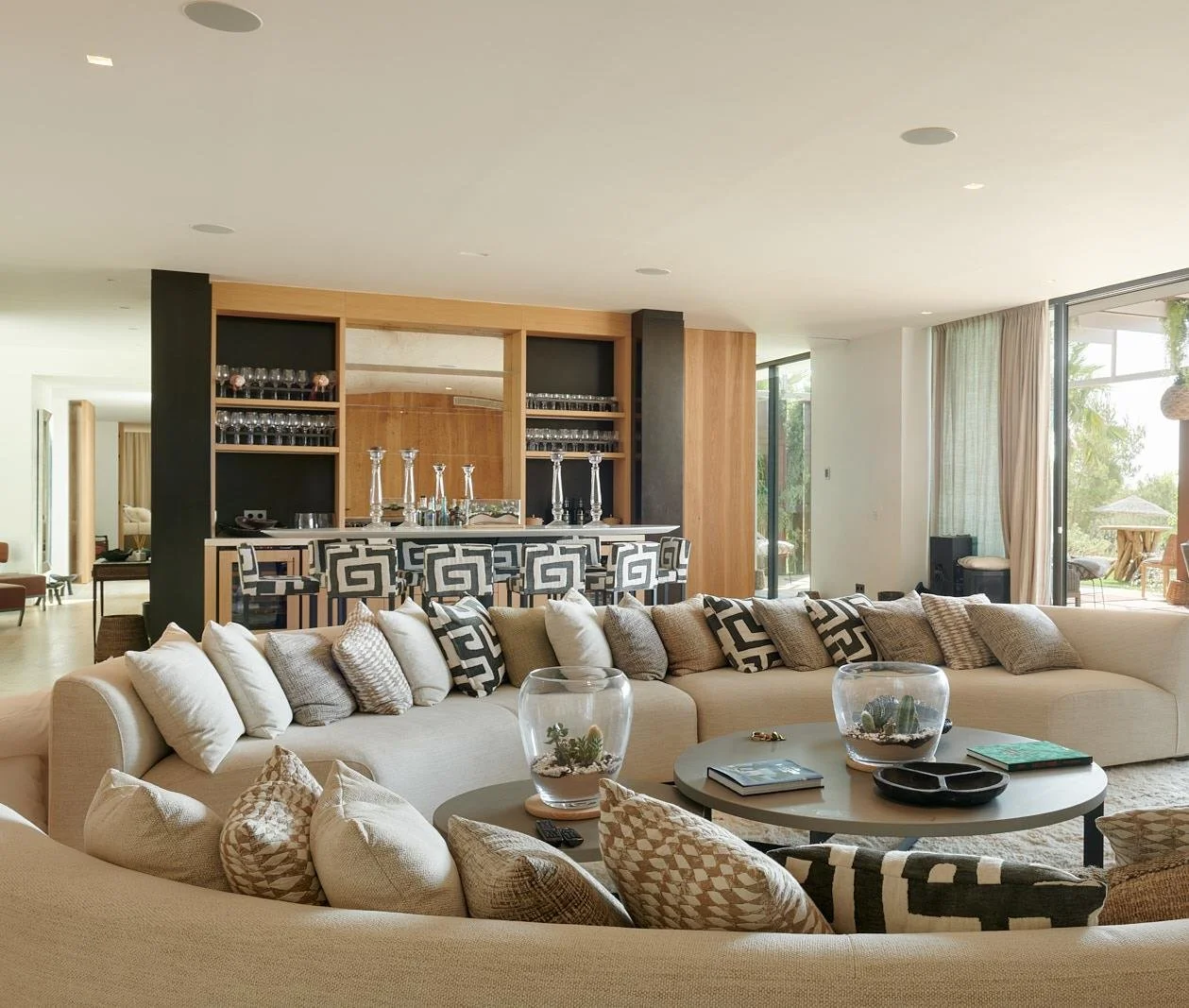 Modern living room with beige sectional sofa, patterned pillows, glass terrariums on coffee tables, open kitchen with bar area, and large windows with outdoor view.