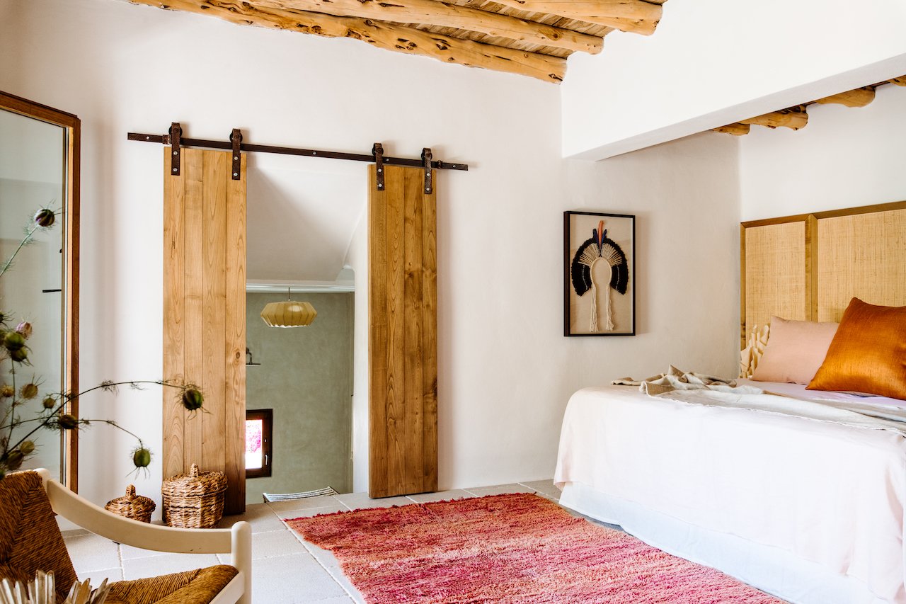 A bedroom with white walls, a bed with beige and bronze pillows, a wooden headboard, a colorful red rug, a sliding barn door, a mirror, and wall art depicting a Native American headdress.