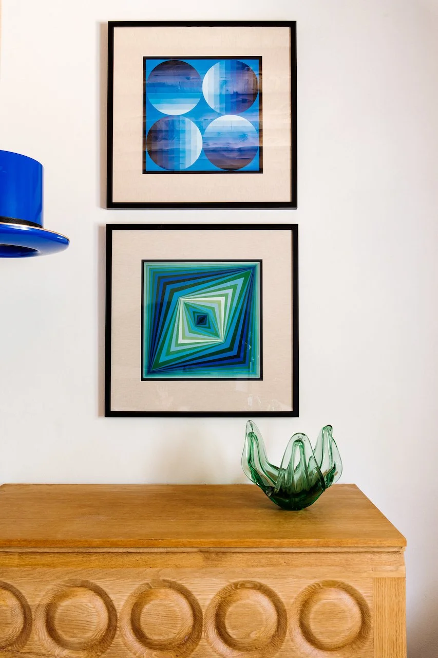 Two framed abstract geometric artworks on a white wall above a wooden sideboard, with a green glass vase on top.