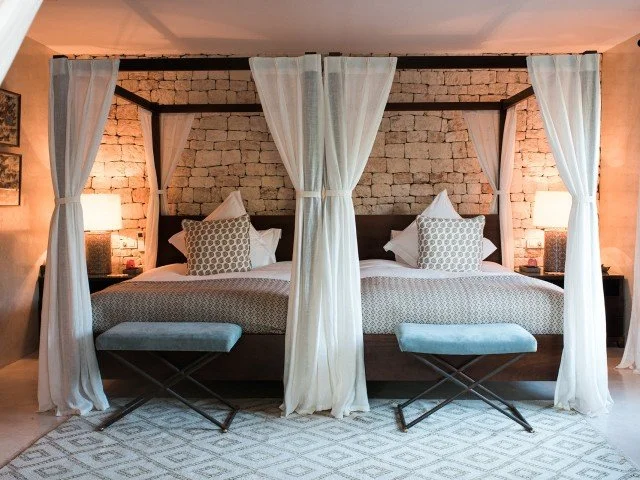 A bedroom with a four-poster bed, white curtains, and a brick accent wall. Two nightstands with lamps, and two upholstered benches at the foot of the bed.