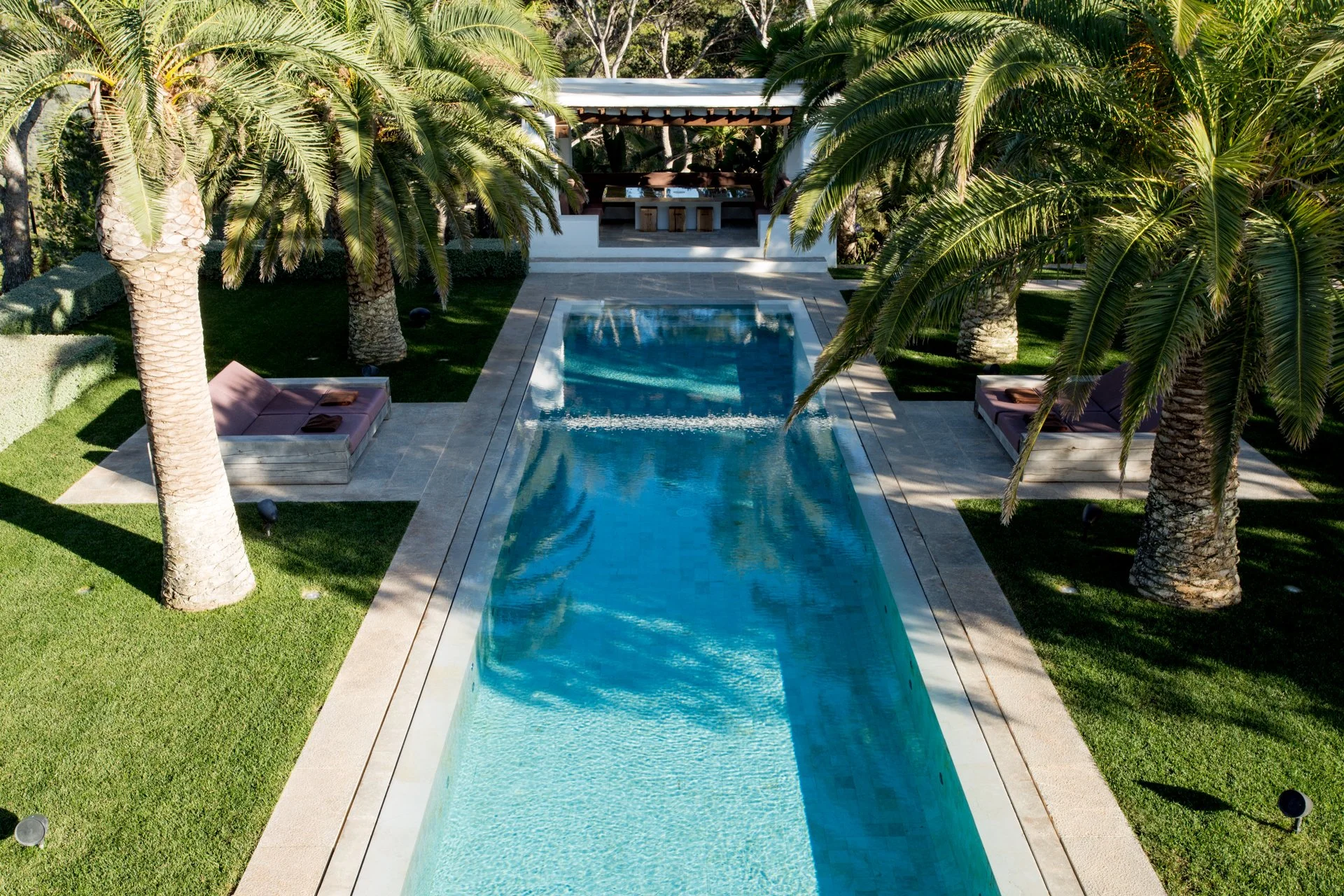 A rectangular swimming pool surrounded by green grass and tall palm trees. There is a shaded seating area with a table and stools in the background.