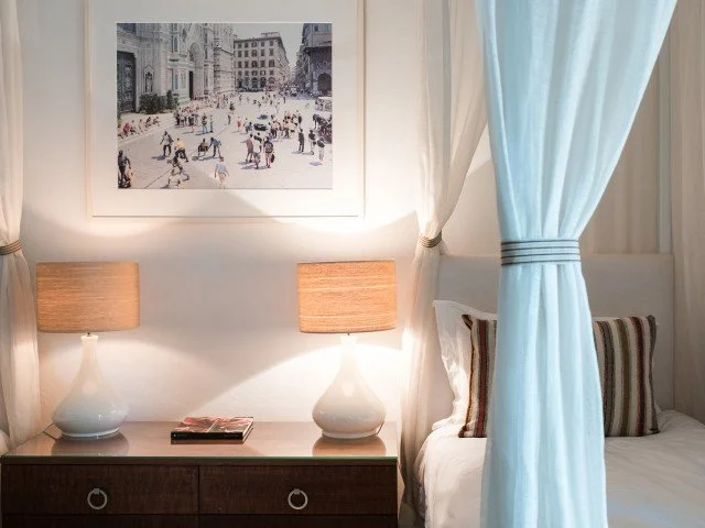 Interior of a bedroom featuring a nightstand with two white table lamps, a stack of magazines, a bed with striped pillows, and a blue curtain tied back to reveal a white canopy.