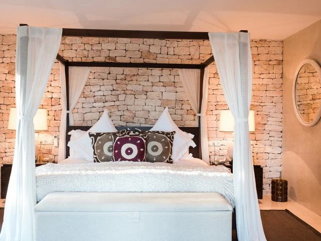A bedroom with a white canopy bed, white curtains, stone brick wall, and two bedside lamps with beige shades.