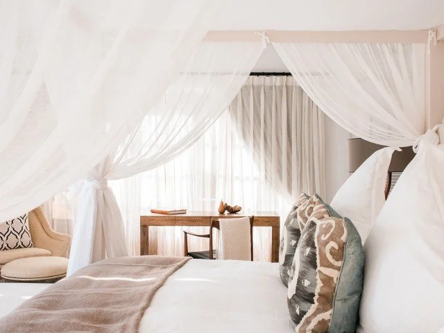 Bedroom with four-poster bed draped with white curtains, decorative pillows, wooden desk with a chair, window with white curtains, and armchair with patterned cushions.