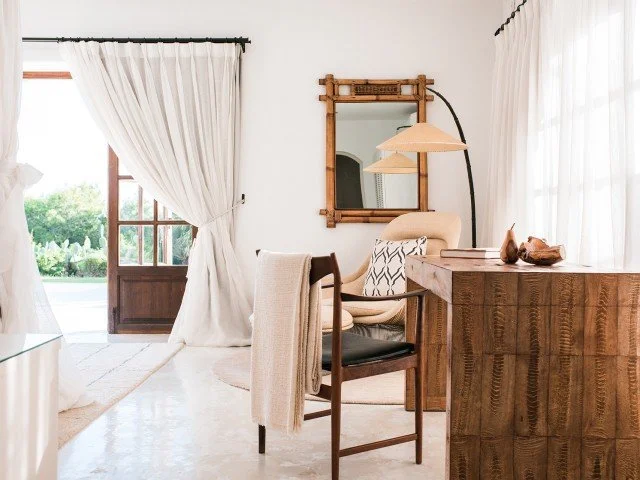 Cozy living room with a wooden desk, two chairs, a large mirror, a floor lamp, and open curtains revealing a balcony view of lush greenery.