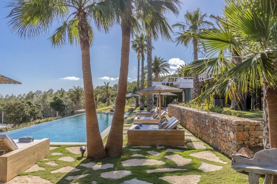 View of a luxurious outdoor pool area with swimming pool, lounge chairs with towels, palm trees, and a stone wall, under a bright blue sky with some clouds.