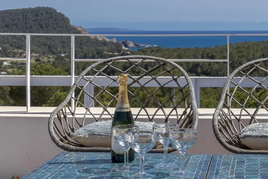 A table set with a champagne bottle and glasses, on a balcony overlooking a scenic landscape with hills and the ocean in the background.