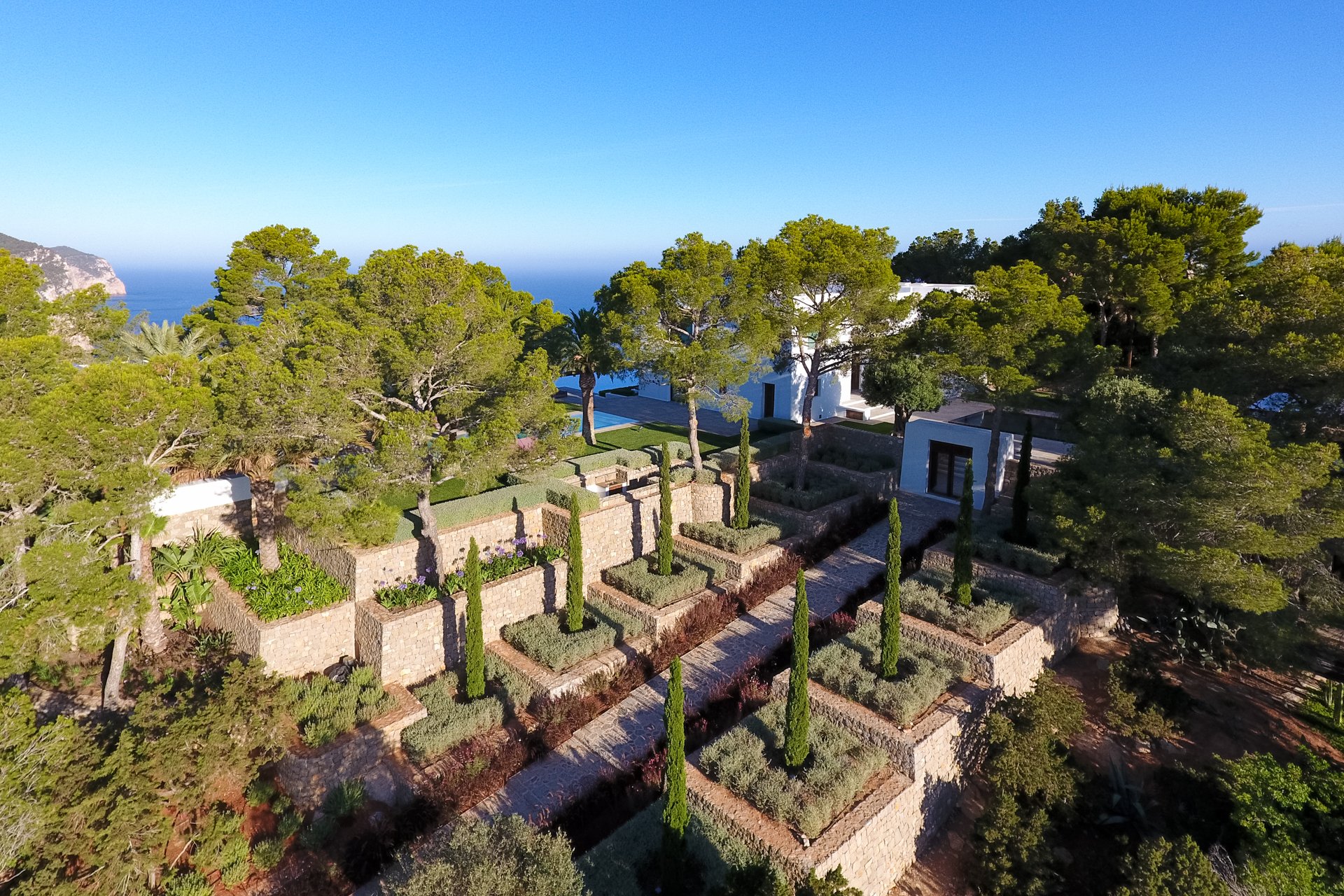 Aerial view of a modern white house among trees, with a terraced garden and stone walkway, ocean and mountains in the background.