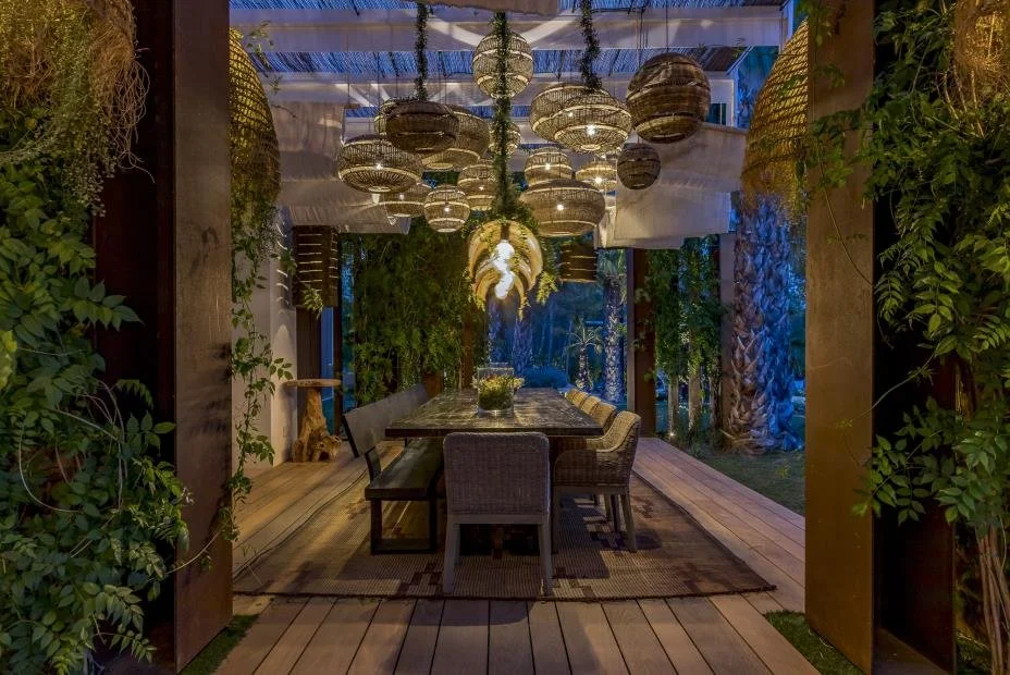 Outdoor dining area with a long table, chairs, hanging lanterns, and greenery.