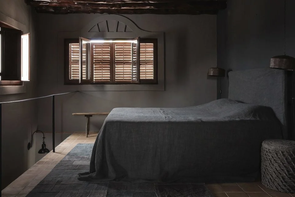 A bedroom with dark walls, a bed with a gray bedspread, wooden shutters on the windows, and minimal decor.