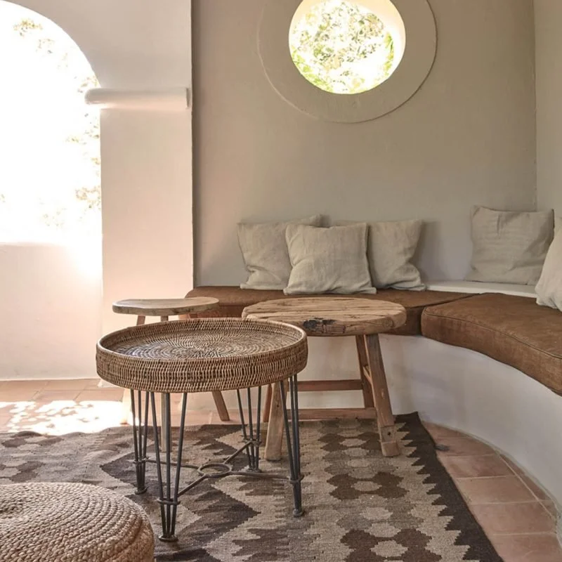 Cozy indoor sitting area with a curved built-in sofa adorned with beige cushions, wooden and woven side tables, a patterned rug, and a small circular window with views of greenery outside.